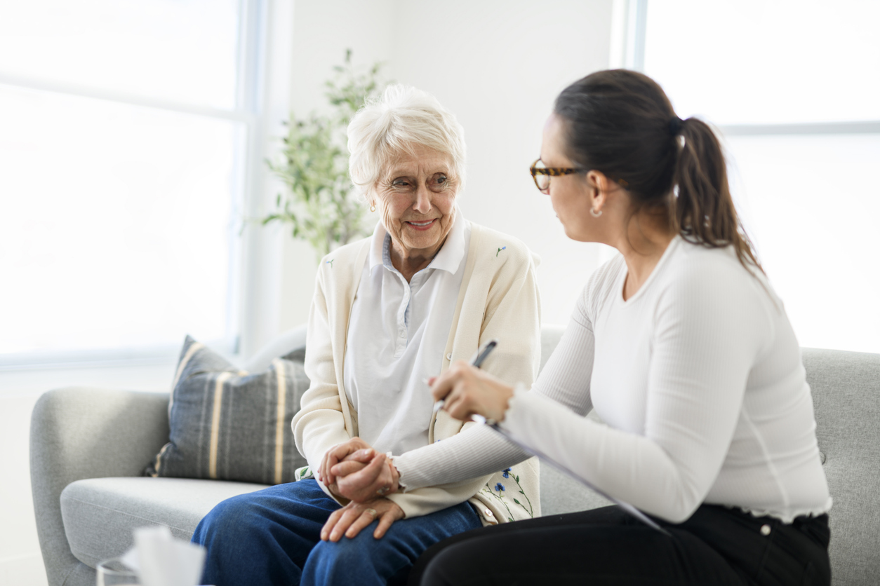 An elderly woman on a gray sofa smiles while a younger woman with a ponytail and glasses talks to her, holding a clipboard.